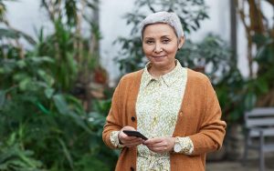 senior woman standing outside holding mobile phone and smiling, plants and greenery behind her, lady wears mustard cardigan and cream blouse