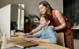 young boy sitting at desk with a computer. His mother stands behind him to help on the computer.