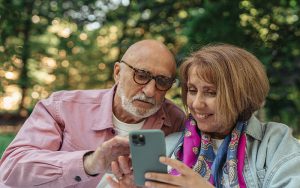 and older man and woman sitting outside surrounded by nature. The woman is holding a mobile phone.