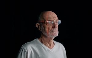 an elderly man stands looking sombre against a black background