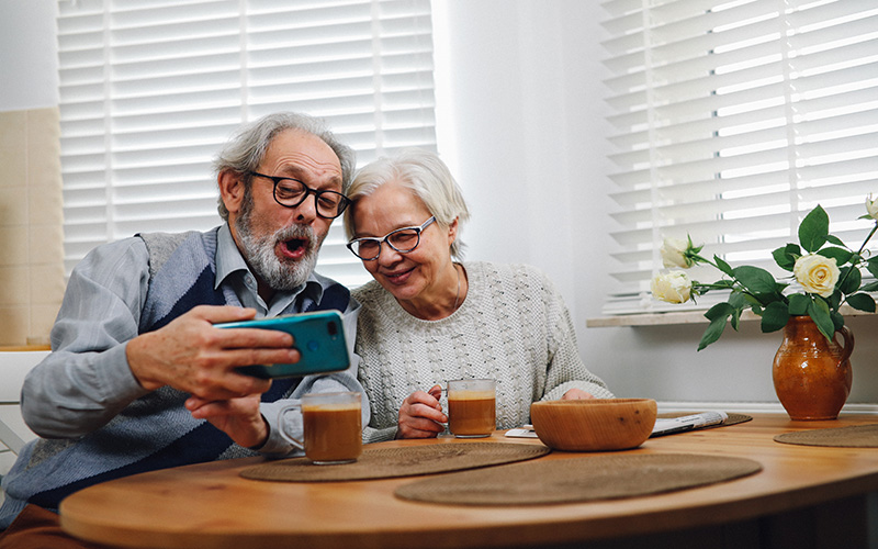 an elderly man and woman sit at a table, the man holds a mobile phone and they're both smiling and laughing