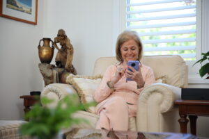 an elderly lady sits on an armchair, she holds a purple phone in her hand and is smiling