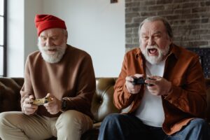 two elderly men sit on a couch playing a console video game, they both hold video game controllers and have animated facial expressions