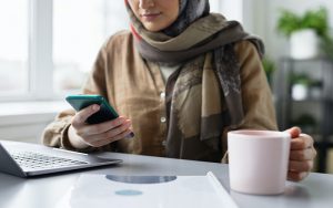 woman sitting at her desk with a phone in her hand and a mug in the other hand. Laptop is on the desk.