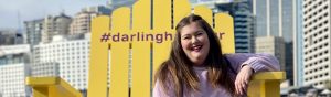 Corinne sits on a large yellow oversized deck chair with Darling Harbour in the background