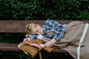 a homeless woman sleeps on a wooden park bench outside with a blanket