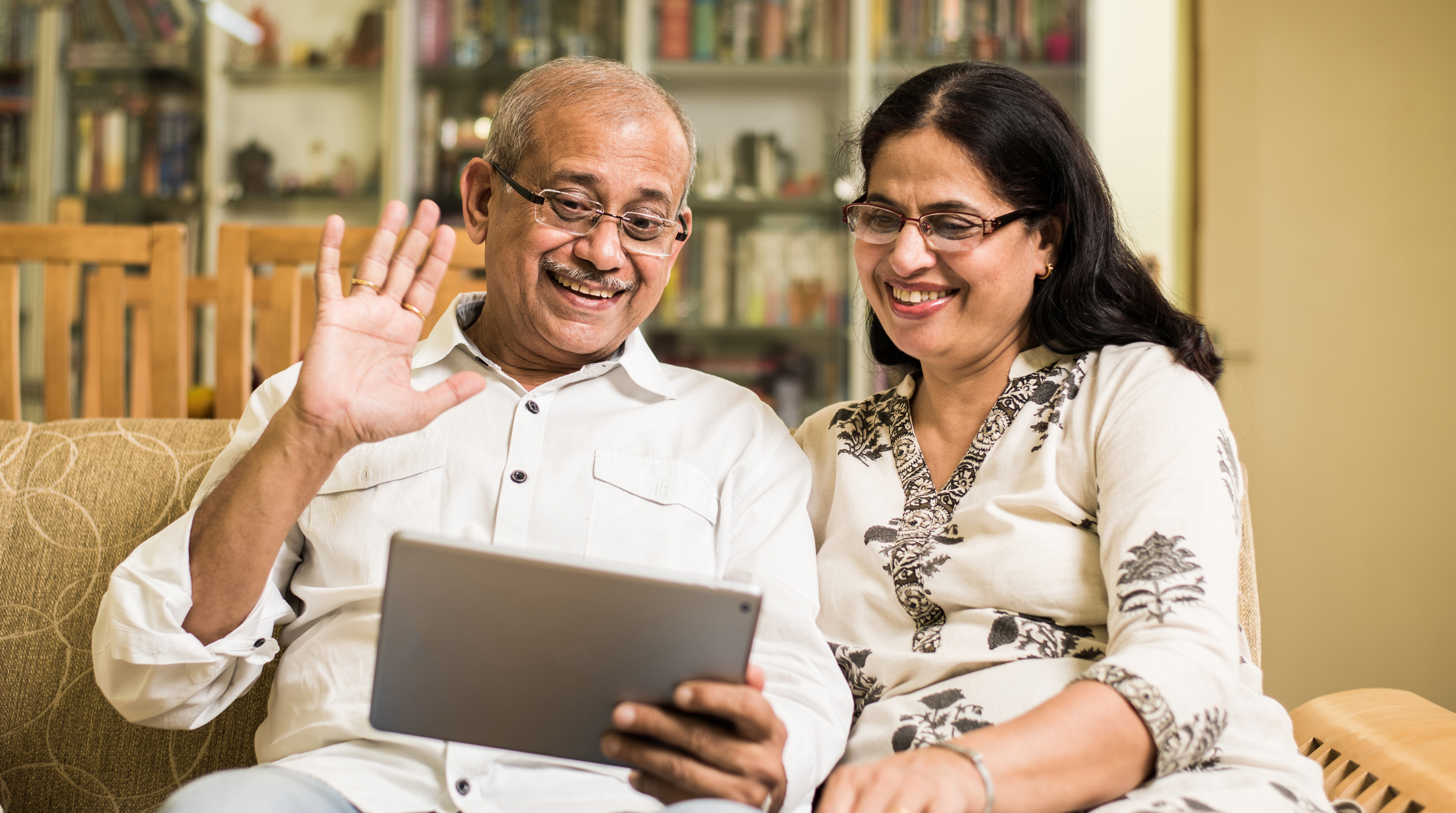 a man and a woman sitting on a couch with an ipad in hand, waving