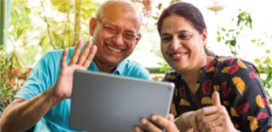 elderly man and woman sitting outside, greenery in background. The man is holding an iPad and he is waving, they are both smiling.