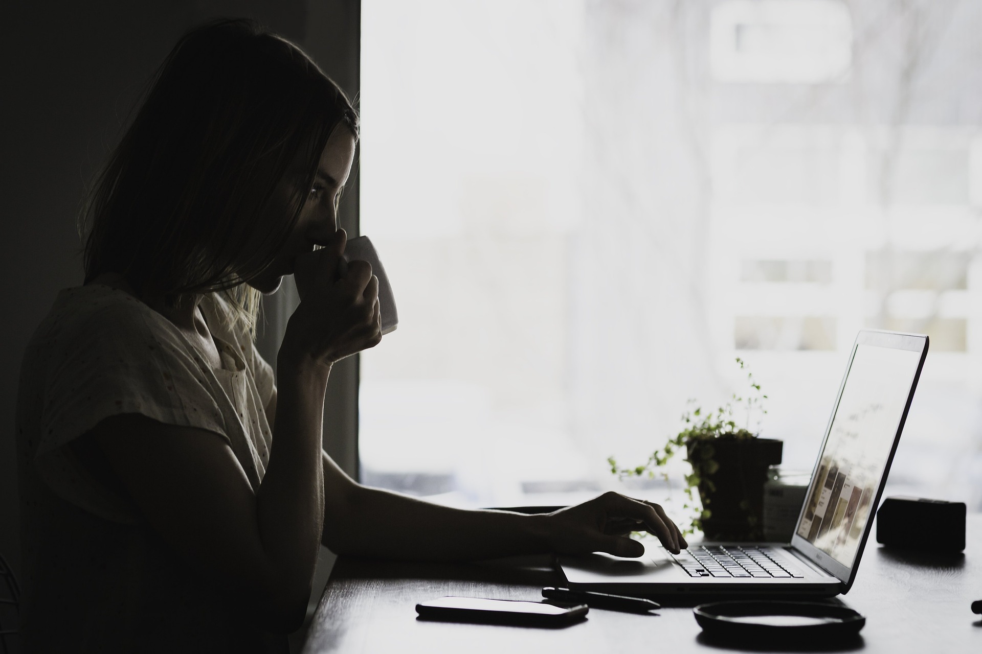 A woman sits at a desk with a laptop, she holds a cup to her mouth which she is drinking