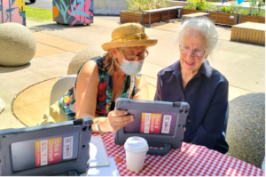 Two women sit at a table outside. On the table is and iPad which the two women are looking at. In the background are some plants.