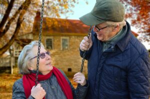 An elderly woman sits on a swing outside with an elderly man standing beside her. There is a brick building behind them and a tree with orange leaves.