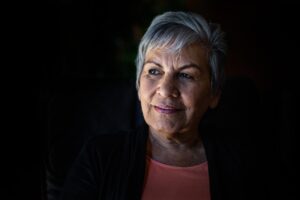 An elderly woman stands looking to her left with a slight smile on her face against a dark background.